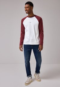 Camicia bianca a maniche lunghe con maniche raglan bordeaux, caratterizzata da un piccolo dettaglio testuale. Indossata con jeans blu e scarpe da ginnastica beige.