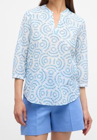 Blusa trasparente con un motivo a spirale tie-dye blu, colletto alla mandarino e maniche a tre quarti, abbinata a pantaloni corti azzurri.