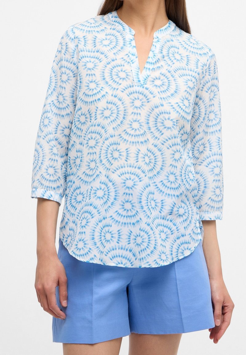 Blusa trasparente con un motivo a spirale tie-dye blu, colletto alla mandarino e maniche a tre quarti, abbinata a pantaloni corti azzurri.