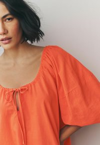 Pomarańczowa bluzka z krótkim rękawem, o szerokim dekolcie i sznurkiem na przodzie. Teksturyzowana tkanina o lekkości.