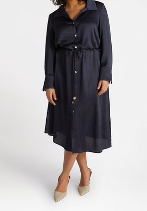 Frau trägt ein marineblaues Midikleid mit langen Ärmeln, Knopfleiste, dünnen vertikalen Streifen und beigefarbene Slingbacks mit spitzem Zehenbereich.