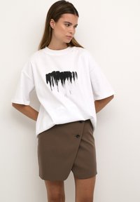 Tricou alb supradimensionat cu imprimeu din pensulă neagră, asortat cu o fustă înfășurată maro, dotată cu două nasturi și o textură subtilă.
