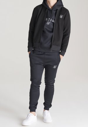 Sort atletisk træningsdragt med lynlåstrøje, hættetrøje og tilspidsede bukser. Blødt stof med logoaccenter. Kombineret med hvide sneakers.