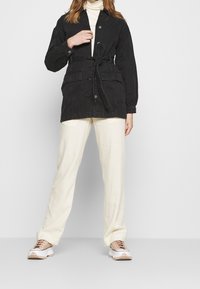 Giacca in velluto nero con vita cinturata, chiusura a bottoni e due tasche frontali, indossata su un leggero dolcevita e pantaloni beige.