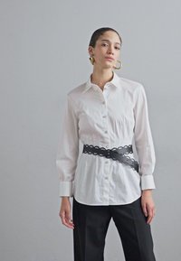 White long-sleeve shirt with a button-down front, paired with a black sculpted belt. The fabric is smooth, and the shirt features a classic collar.