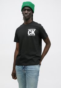 Czarny bawełniany t-shirt z białym nadrukiem z napisem "Calvin Klein" i "Established 1968", zestawiony z jasnoniebieskimi jeansami i zieloną czapką beanie.
