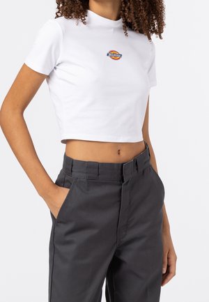 Woman wearing a white cropped Dickies t-shirt and high-waist dark gray pants, with one hand in pocket against a white background.