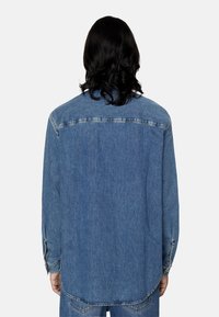 Camicia in denim, blu medio, maniche lunghe, polsini a bottone, dettagli sulla spalla posteriore, orlo curvo, vestibilità casual, tessuto testurizzato, senza motivi visibili.