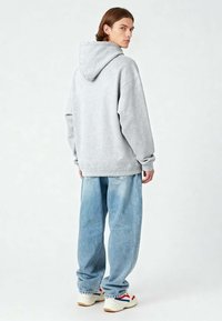Felpa grigia chiara con cappuccio regolabile, vestibilità oversize, polsini a coste, abbinata a jeans azzurri larghi e sneaker bianche.