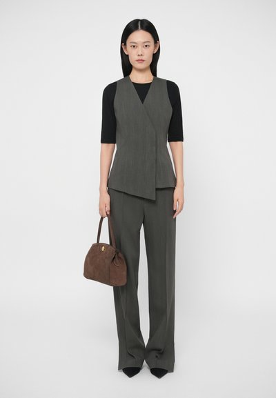 Γυναίκα ντυμένη με γκρι αμάνικο σακάκι και ταιριαστά παντελόνια πάνω από μαύρο τοπ, κρατώντας καφέ τσάντα, στέκεται μπροστά σε λευκό φόντο.