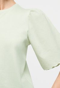 Ανοιχτό πράσινο τοπ με κοντά μανίκια φουσκωτά και κυματιστά τελειώματα στα μανίκια, φορεμένο από άτομο σε λευκό φόντο.