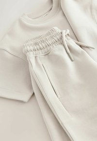 Lysebeige sweatshirt- og joggingtøjssæt lavet af blødt stof. Joggingbukserne har elastisk linning og sidelommer samt en afslappet pasform.