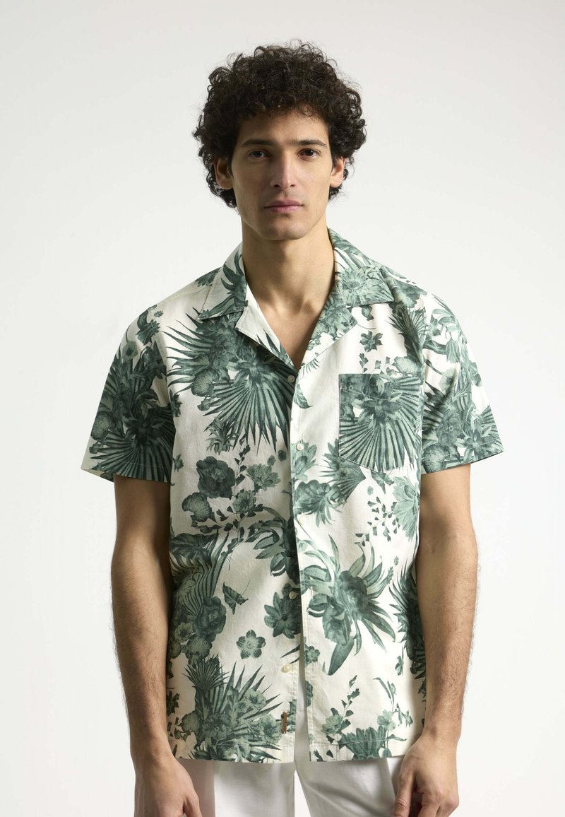 Camisa de manga corta en tela crema con patrones florales tropicales verdes y un bolsillo en el pecho. Presenta un cuello clásico y botones.