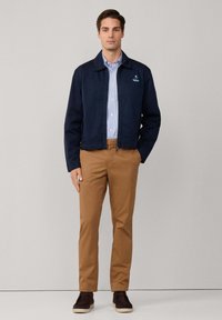 Giacca navy con colletto, zipper frontale e logo ricamato; camicia a righe con bottoni; pantaloni marroni a gamba affusolata; scarpe scure con suole chiare.