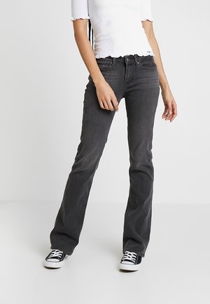 Person wearing black fitted bootcut jeans, black and white sneakers, and a white short-sleeve ruffled top, standing against a plain background.