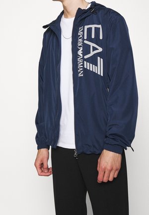 Mężczyzna ubrany w granatową kurtkę na zamek EA7 Emporio Armani na białej koszuli i czarnych spodniach, stojący na jednolitym tle.