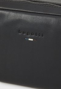 Borsa in pelle nera con logo embossato "bugatti" e un piccolo accento colorato di blu, bianco e marrone scuro vicino al logo. Chiusura con cerniera.