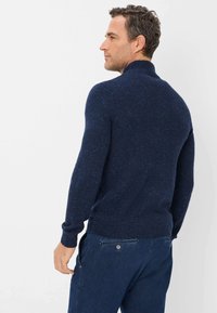 Navyblauer Strickpullover mit strukturiertem Muster, hohem Kragen und gerippten Bündchen, kombiniert mit dunklen Jeans, von hinten gezeigt.