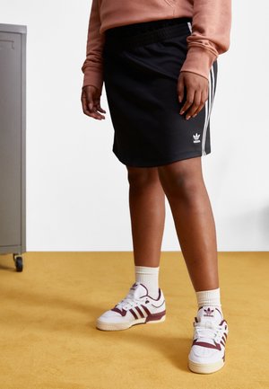 Personne portant une jupe noire Adidas avec des rayures blanches et un logo, des baskets blanches Adidas avec des détails bordeaux, debout sur un sol moutarde.