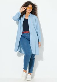 Abito chemisier in denim azzurro chiaro con maniche lunghe, frontale con bottoni e orlo asimmetrico. Abbinato a jeans a pois blu scuro e sneaker bianche.