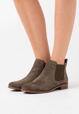 Benen met bruine suède Chelsea-laarzen met elastische zijkanten en lage gestapelde hakken op een witte achtergrond.