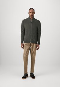 Dark green ribbed zip-up sweater with a high collar, paired with beige trousers and black sneakers featuring brown soles.