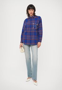 Blåternet skjorte med lange ærmer, knaplukning foran og brystlomme. Båret med lyseblå jeans og hvide spidse flade sko.