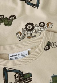 Beige børnetrøje med trykte illustrationer af entreprenørmaskiner, herunder gravemaskiner og læssemaskiner.