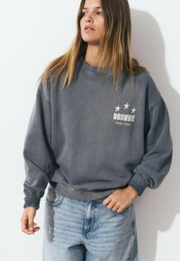 Joven mujer con cabello largo, vistiendo un sudadera gris desvaído de "Brownie World Tour" y jeans de mezclilla azul claro, mirando hacia un lado.