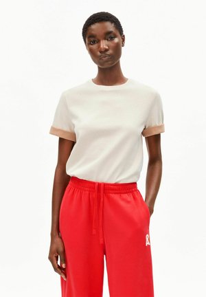 T-shirt beige clair avec des poignets courts marron, associée à un pantalon de survêtement rouge vif présentant un motif blanc sur la jambe gauche.