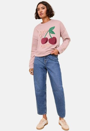 Un sweat-shirt rose avec des cerises brodées et du texte, associé à un jean bleu ample et des baskets beige clair. Style décontracté, coupe relax.
