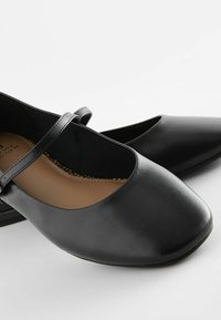 Brogue in pelle nera con punta arrotondata, cinturino singolo sul collo del piede e suola piatta. L'interno presenta un morbido rivestimento beige.