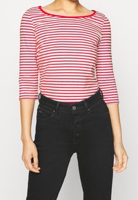 Rot-weiß gestreiftes Langarmshirt mit weitem Ausschnitt, kombiniert mit hochgeschnittenen schwarzen Jeans, die über einen Knopfverschluss und fünf Taschen verfügen.