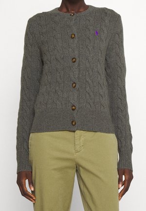 Cárdigan gris oscuro de punto con botones de carey y pequeño logo bordado en púrpura en el pecho, combinado con pantalones en tono oliva claro.