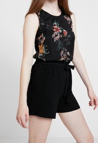 Černý květinový bezrukávový top s barevnými květy, v kombinaci s černými volnými šortkami se šňůrkovým páskem v pase.