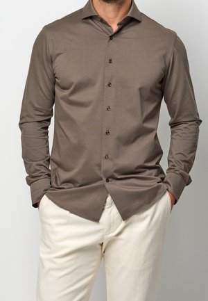 Uomo che indossa una camicia marrone aderente a maniche lunghe con colletto e pantaloni color crema, con le mani infilate casualmente nelle tasche.