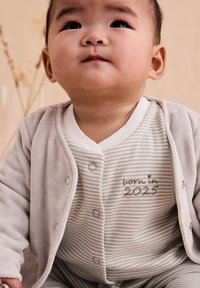 Ljusgrå plyschjacka över en randig grå och vit jumpsuit med knappar, med "född 2025" broderat med grå tråd.