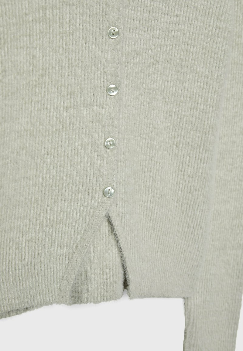 Weicher, hellgrauer, gerippter Strickpullover mit fünf kleinen, durchsichtigen Knöpfen und einem Schlitz am vorderen Saum.