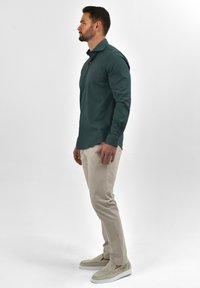 Donkergroen button-up shirt met kraag, gedragen met beige broek en lichtgrijze instapschoenen, met een gladde textuur en een getailleerd ontwerp.