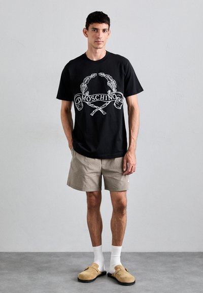 Μαύρο βαμβακερό μπλουζάκι με λευκό γραφικό λογότυπο και σχέδιο. Συνδυασμένο με μπεζ καρό σορτς, λευκές κάλτσες και καφέ slip-on παπούτσια.