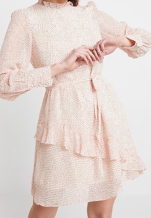 Kobieta w jasno-różowej, długorękawowej sukience w drobne ciemne kropki, z falbaną u dołu i asymetryczną, warstwową spódnicą, pozująca z rękami przy talii.