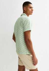 Långärmad skjorta i ljusgrön tyg med ett upprepat vitt geometriskt mönster. Krage design med en avslappnad passform, ihopparad med beiga shorts.