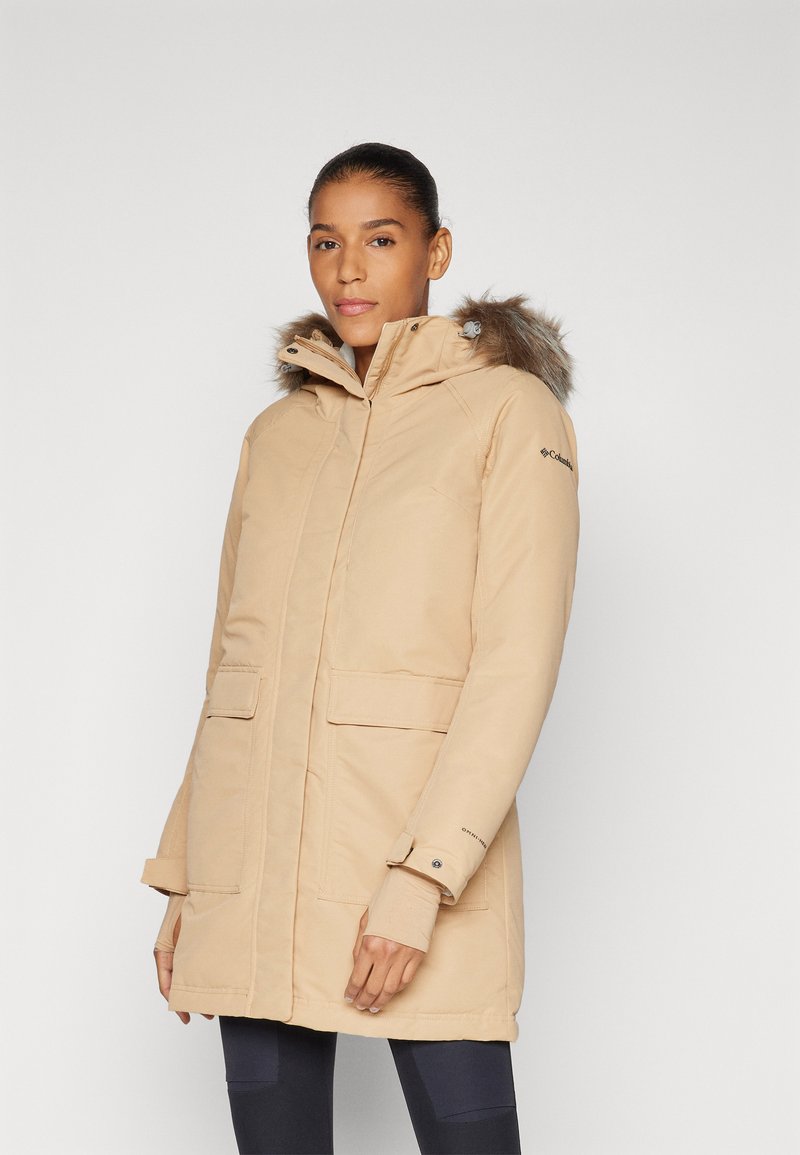 Parka d'hiver beige avec capuche dotée d'une finition en fausse fourrure, fermeture éclair à l'avant, deux grandes poches et poignets avec trou pour le pouce. Tissu lisse et résistant à l'eau.