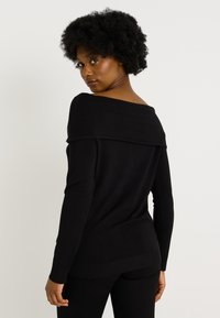 Mujer con peinado afro que lleva un top negro de manga larga y sin hombros, mirando por encima del hombro contra un fondo blanco liso.