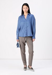 Blue satin shirt with a button-up design, paired with light brown tapered trousers. Model holds a small blue bag, wearing silver shoes.