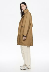 Cappotto oversize marrone con colletto alto, bottoni a pressione e tasche laterali. Abbinato a pantaloni crema e scarpe sportive argentate.