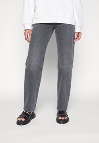 Jeans grigio scuro con vestibilità dritta, realizzati in denim. Abbinati a sandali neri senza lacci con due cinturini. Felpa bianca semplice visibile.
