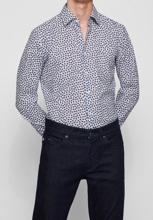 Man draagt een wit overhemd met een donkerblauw bloemenpatroon, ingestopt in een donkerblauwe spijkerbroek. Handen zijn achter zijn rug.