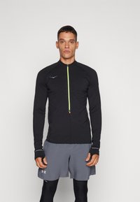 Haut de sport noir à manches longues avec un devant zippé, zip néon contrastant et trous pour les pouces aux poignets, associé à un short gris.
