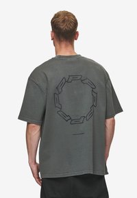 Grå oversized t-shirt lavet af bomuld, med et stort sort cirkelgrafik på ryggen og en rund halsudskæring. Korte ærmer.